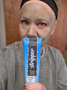 Woman wearing a deep green clay face mask holding a tube of Perfectly Posh The Stripper Deep Cleansing Face and Body Mask with Kaolin, Charcoal, and French Green Clay.
